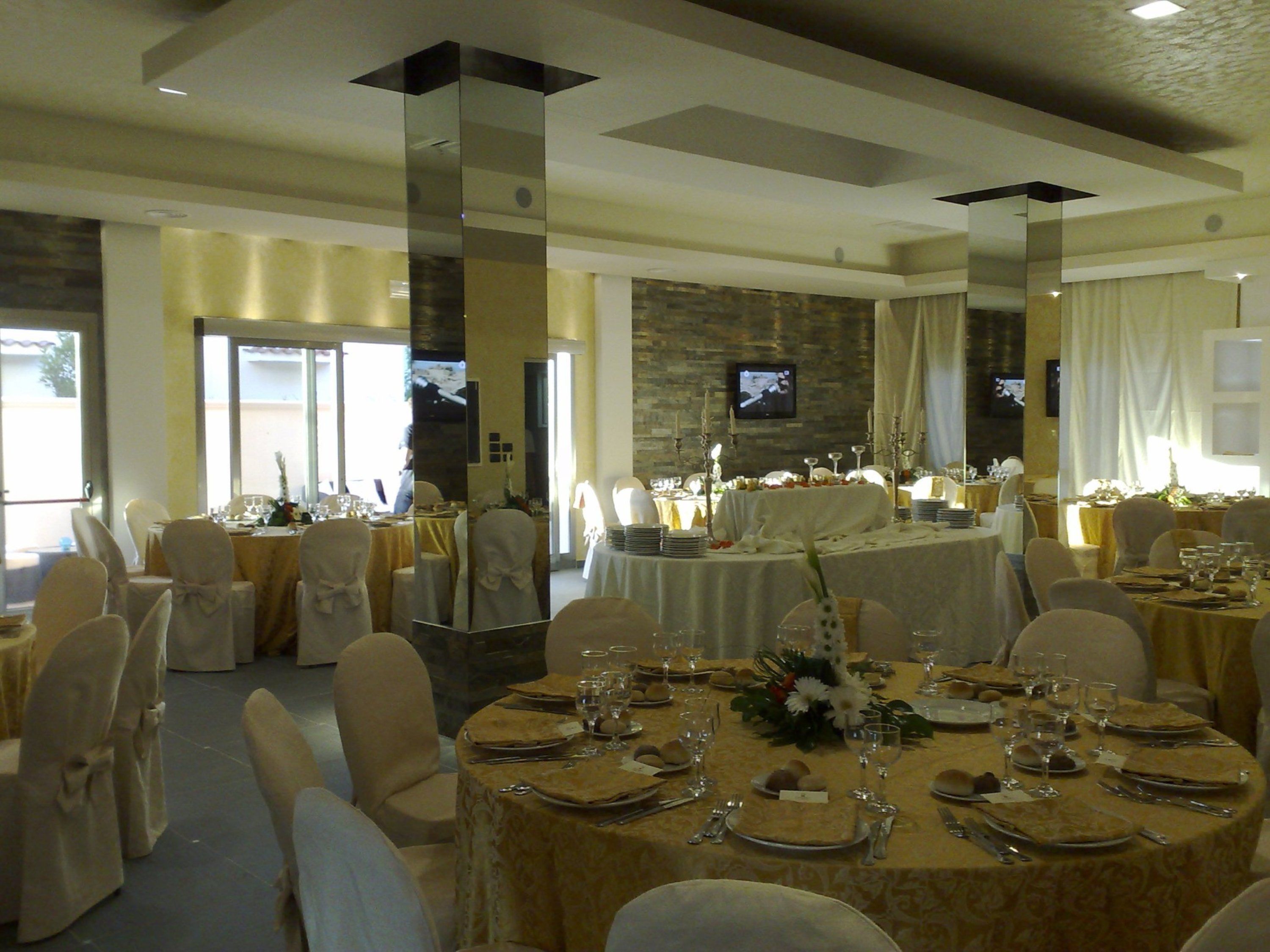 Une salle de banquet avec des tables rondes dressées pour le repas, des chaises recouvertes de housses blanches et un mur de pierre en arrière-plan.