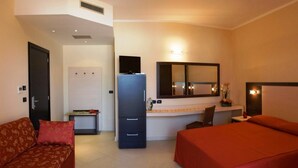 Quarto de hotel com cama, escrivaninha com computador, geladeira, televisão e espelho.