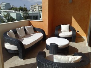 En balkon med fletmøbler, heriblandt en sofa og to stole, samt udsigt til bygninger i baggrunden.