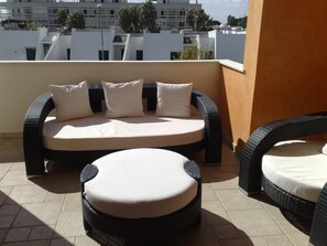 A balcony with a wicker sofa and two round cushions, a tiled floor, and a view of buildings in the background.