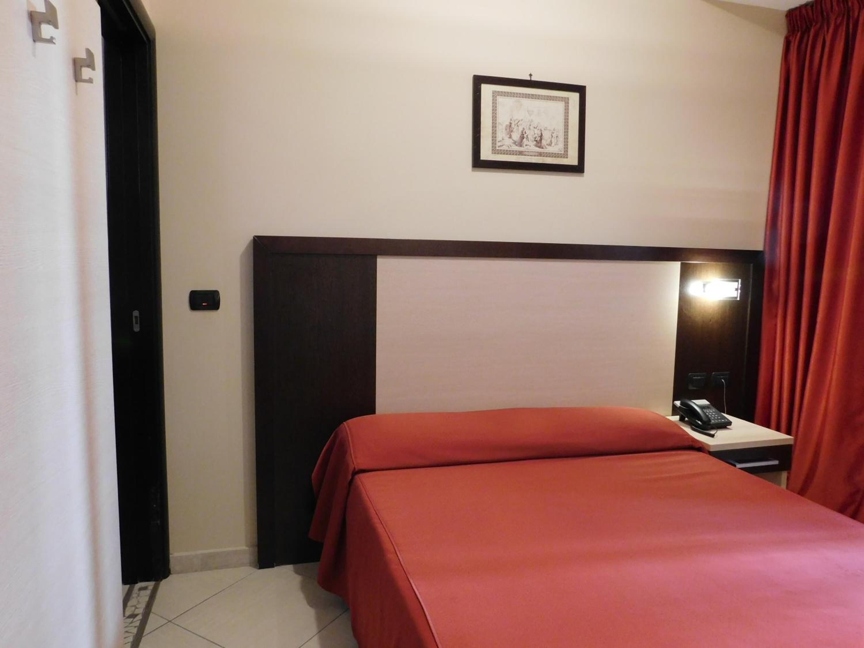 A hotel room with a bed, a bedside table with a phone, a framed picture on the wall, and a red curtain.