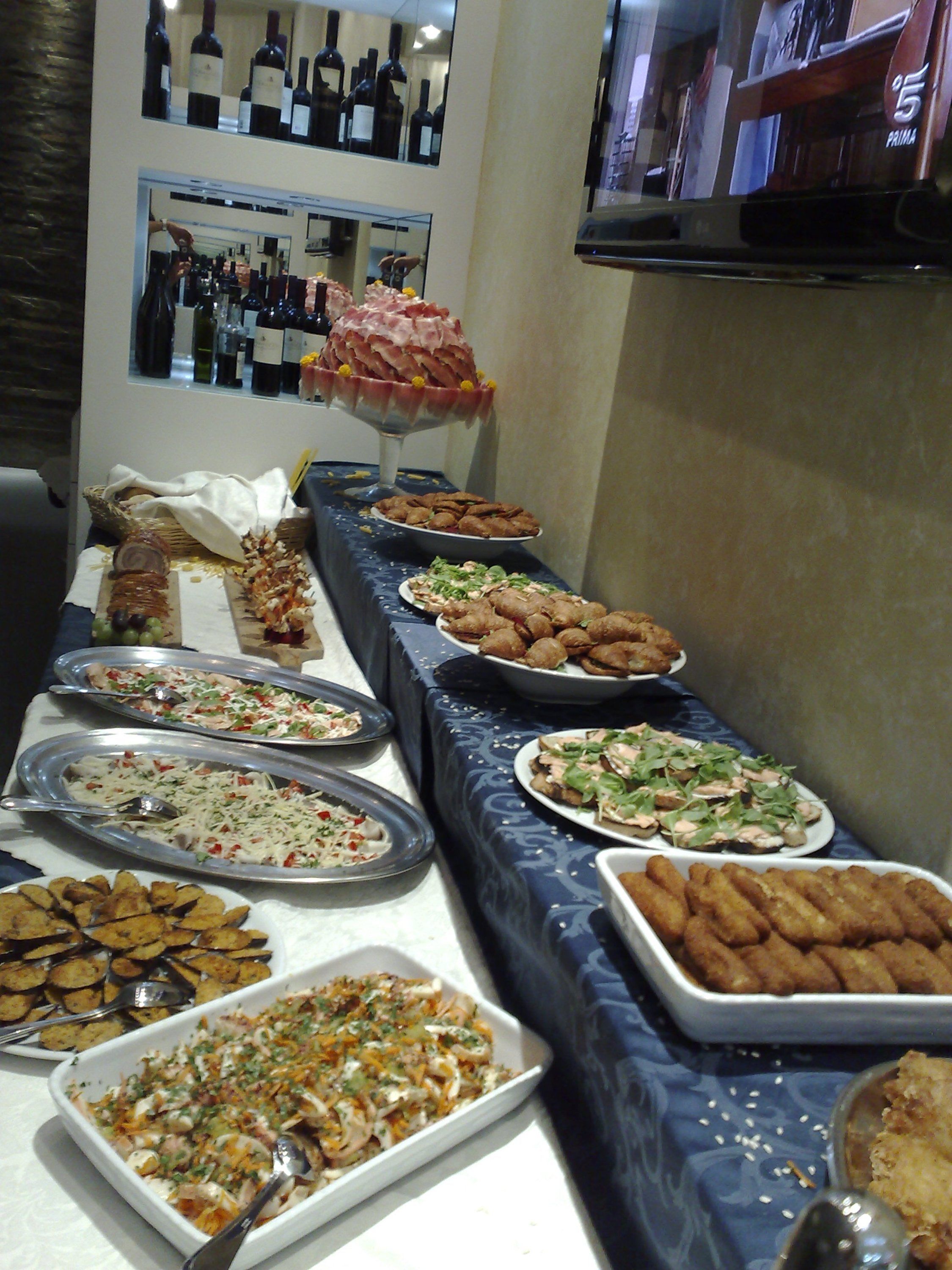 Une table de buffet proposant divers plats, notamment des pâtes, des salades et des plateaux de viande, dans une pièce équipée d’un meuble à vin et d’un téléviseur.