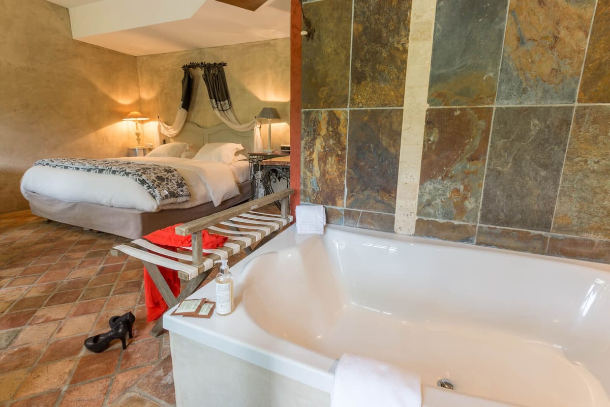 Ein Schlafzimmer mit einer großen weißen Badewanne, einem Bett mit weißer Bettwäsche, einer hölzernen Bank und einer Wand mit Steinfliesen.