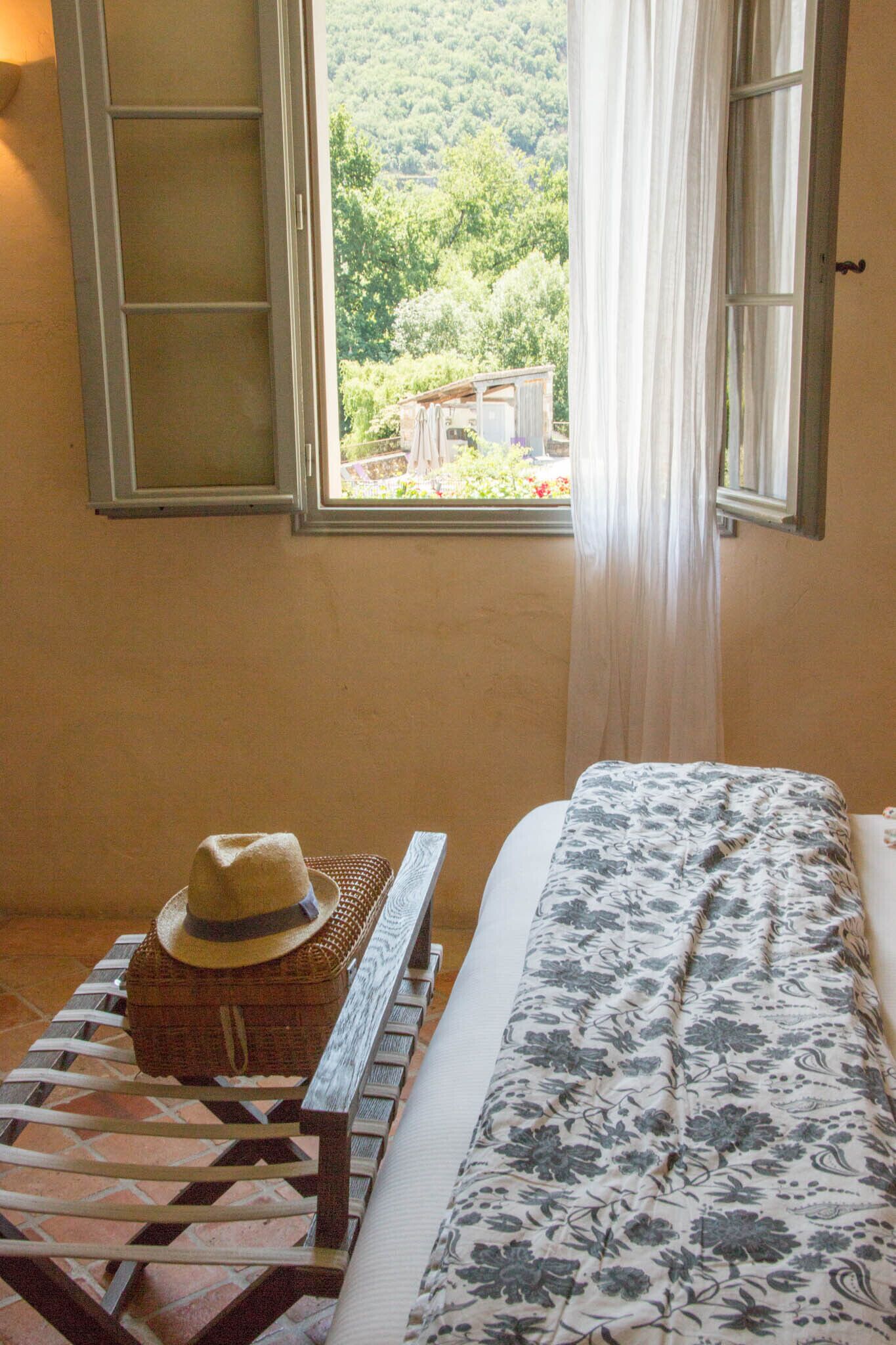 Une pièce avec un lit, une fenêtre avec des rideaux, un chapeau posé sur un banc et une vue sur la verdure à l’extérieur.