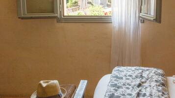 A room with a bed, a window with curtains, a hat on a bench, and a view of greenery outside.
