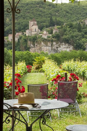 A table with a hat and book, surrounded by chairs, overlooking a lush garden with red flowers and a view of a castle on a cliff.