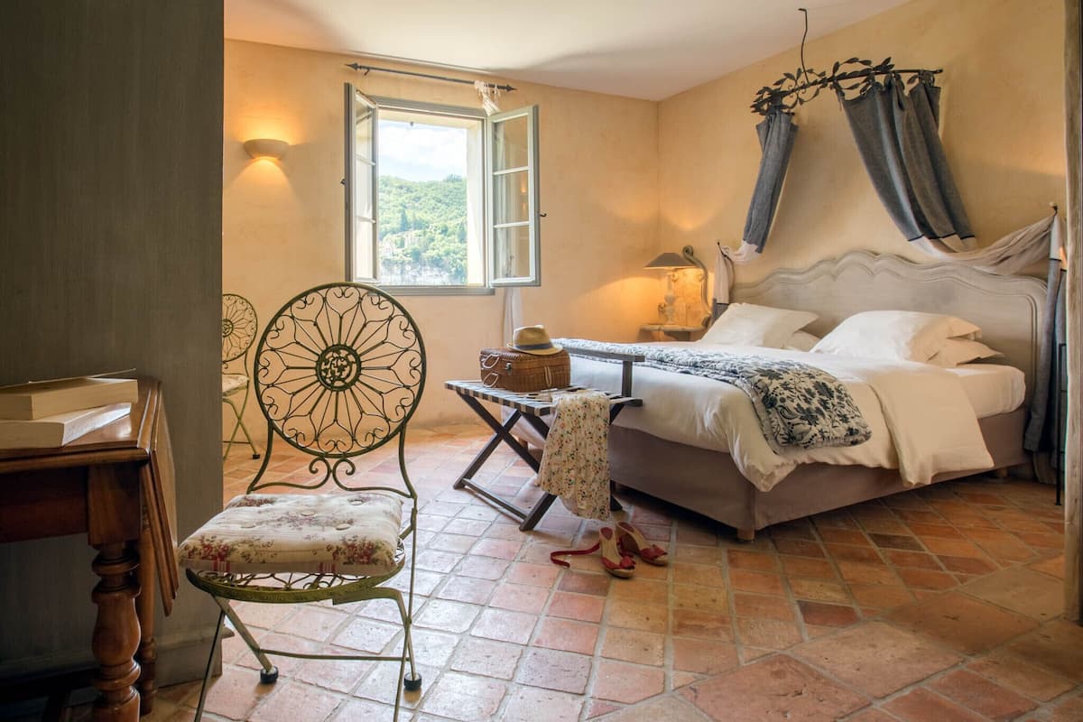 Ein Schlafzimmer mit Bett, Stuhl, Tisch, Fenster und einer Lampe.