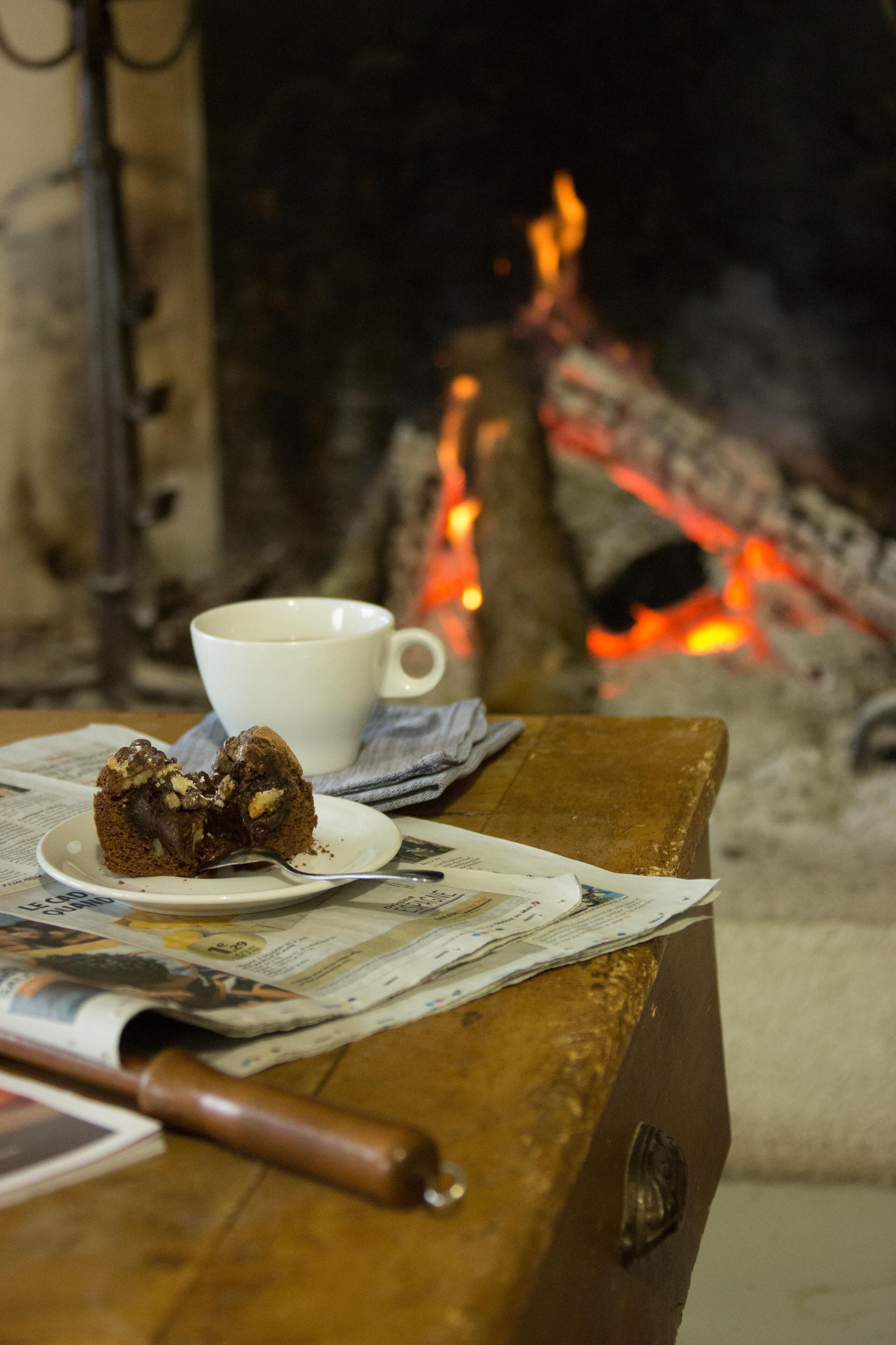 A slice of chocolate cake on a plate with a white cup beside it, placed on a wooden table with newspapers underneath.