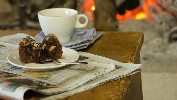 Une part de gâteau au chocolat sur une assiette, à côté d’une tasse blanche, le tout posé sur une table en bois recouverte de journaux.