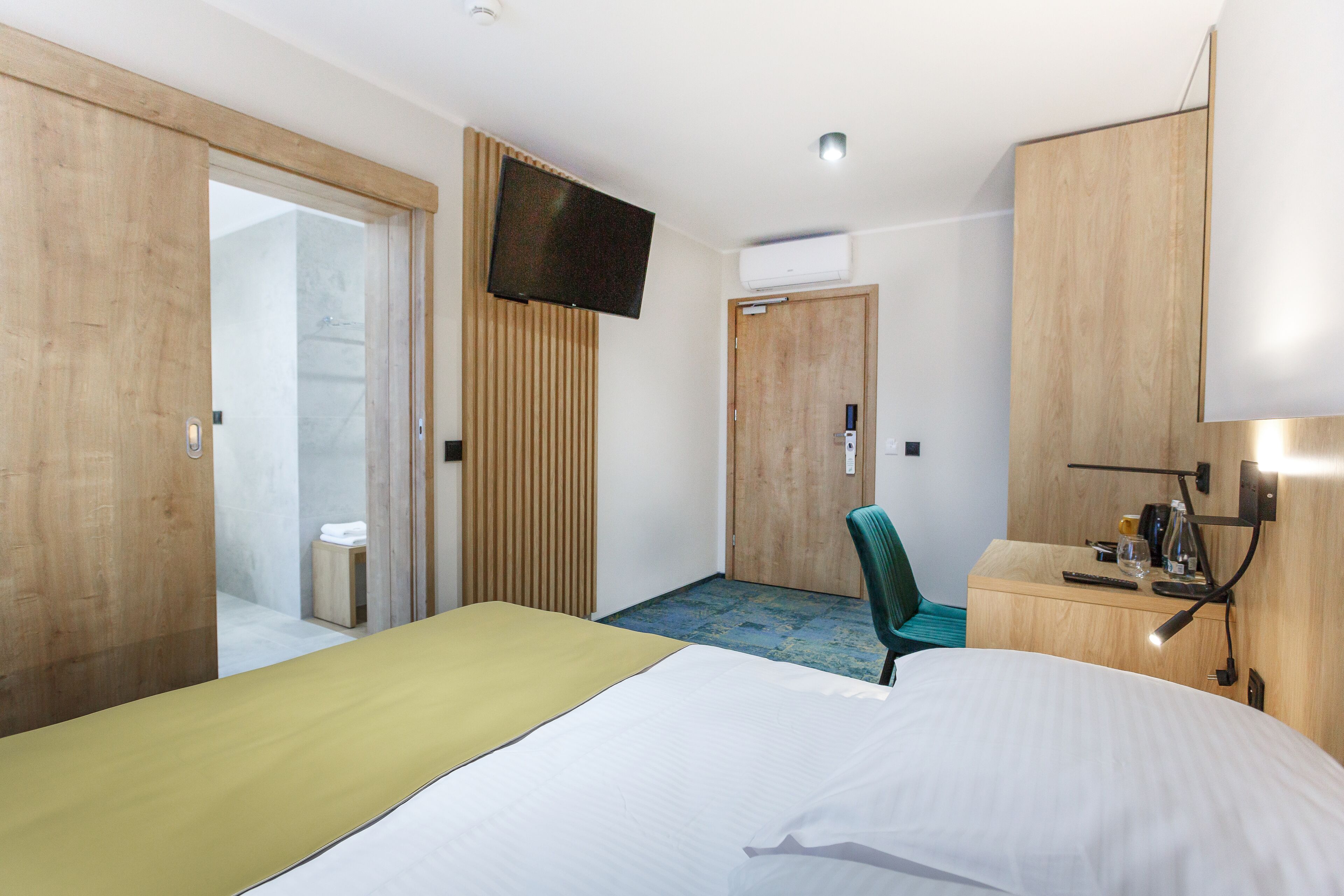 A hotel room with a bed, a TV, a desk with a lamp, and a chair.