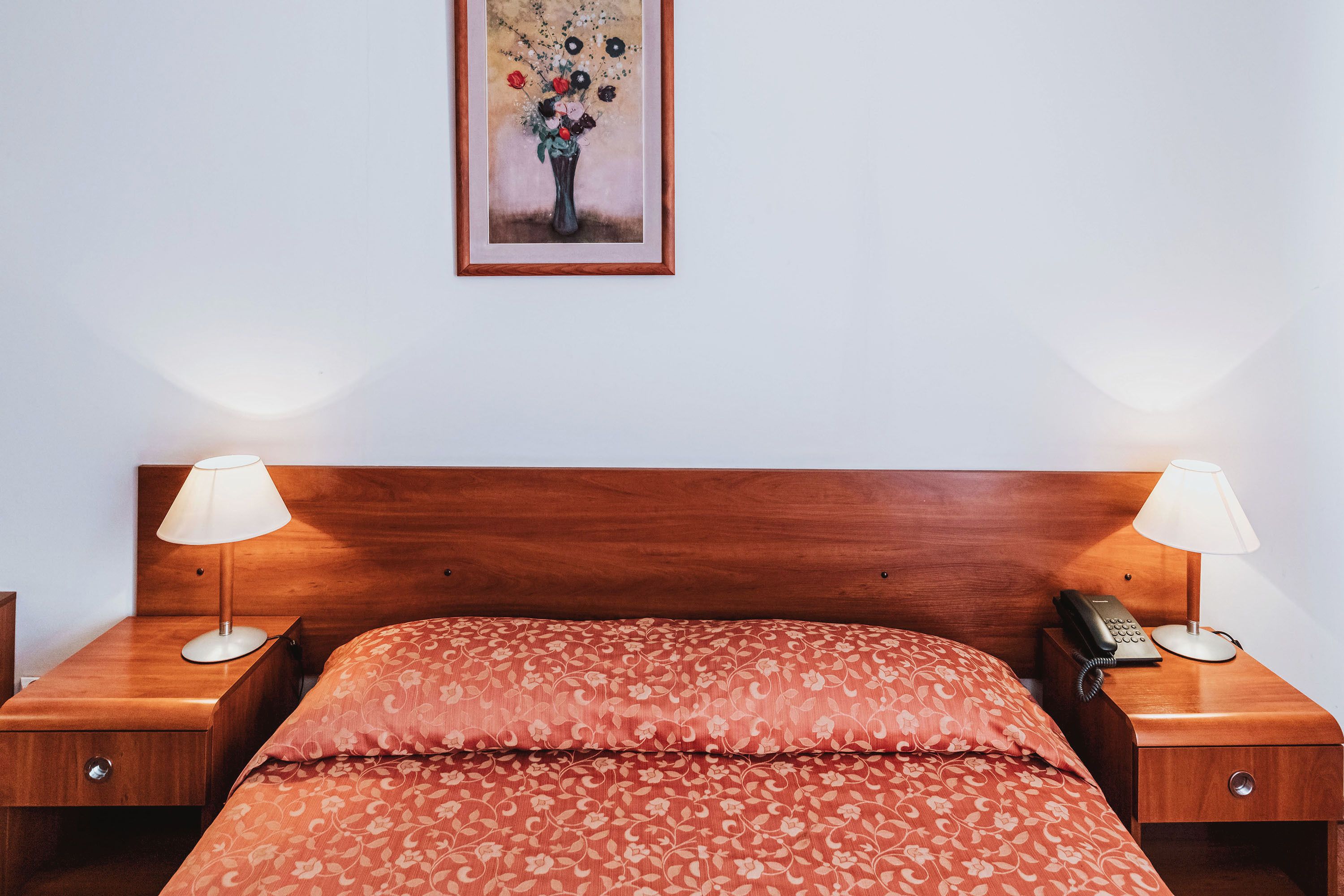 A neatly made bed with floral bedding, two bedside lamps, a nightstand with a telephone, and a framed floral painting on the wall.
