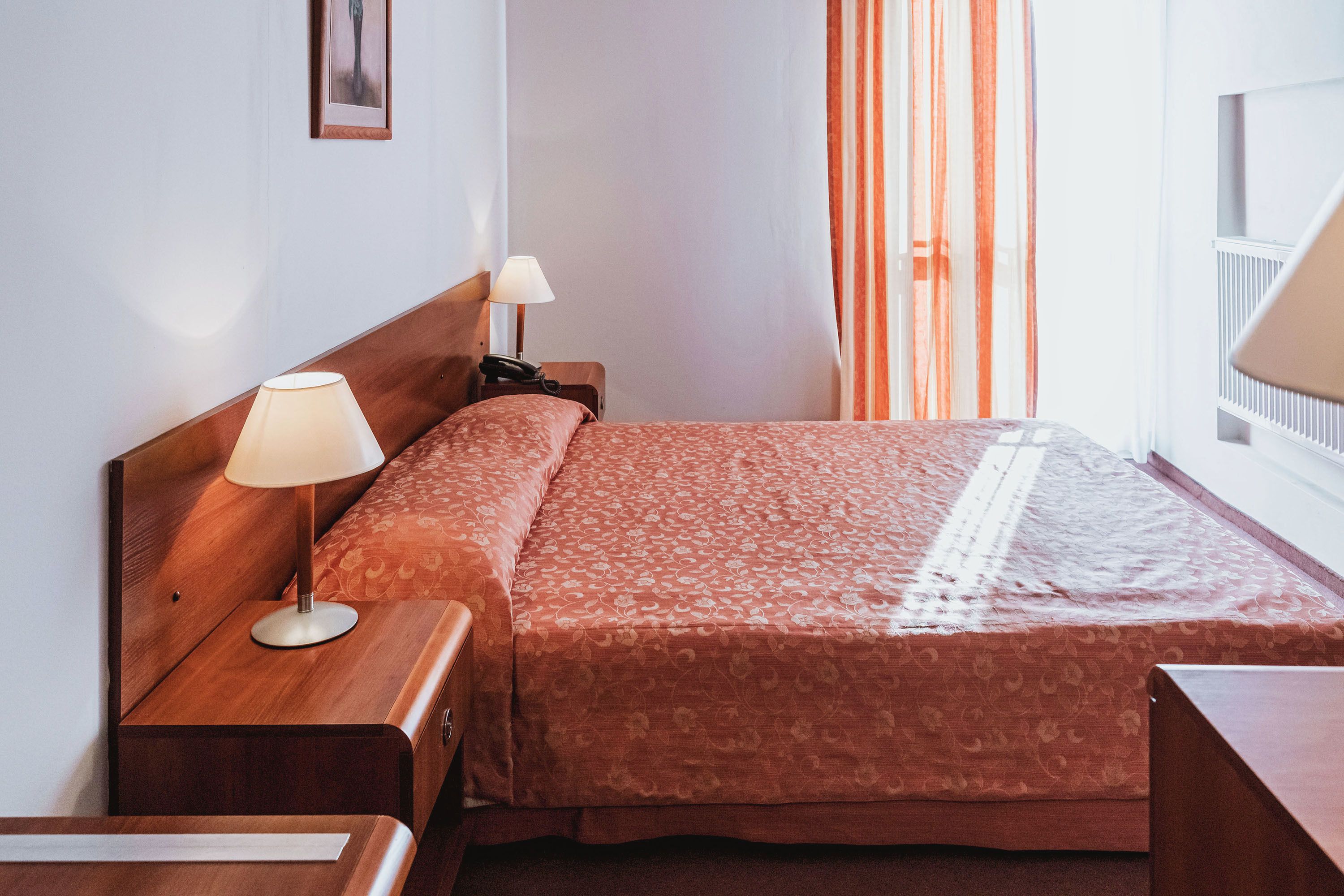 A hotel room with a wooden bed, a bedside table with a lamp, a nightstand, and a window with curtains.