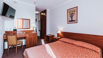 A hotel room with a bed, a desk with a chair, a television, a mirror, a nightstand with a lamp, and a framed picture on the wall.