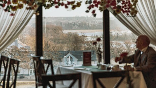 A dining area with a table set for two, a view of a residential area through large windows, and a person standing by the window. - Hotel Gromada Lomza (Lomza)