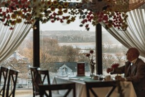 A dining area with a table set for two, a view of a residential area through large windows, and a person standing by the window. - Hotel Gromada Lomza (Lomza)