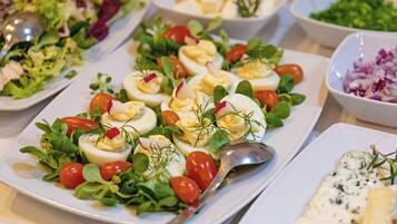 A buffet table with various dishes including a salad with cherry tomatoes, greens, and egg rolls, and a bowl of sliced cheese with herbs.