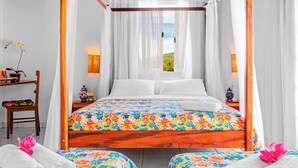 A bedroom with a wooden bed, white bedding, and a colorful floral bedspread. There are two bedside tables with lamps, a chair, and a view of the outdoors. - Villa Baobá (Búzios)