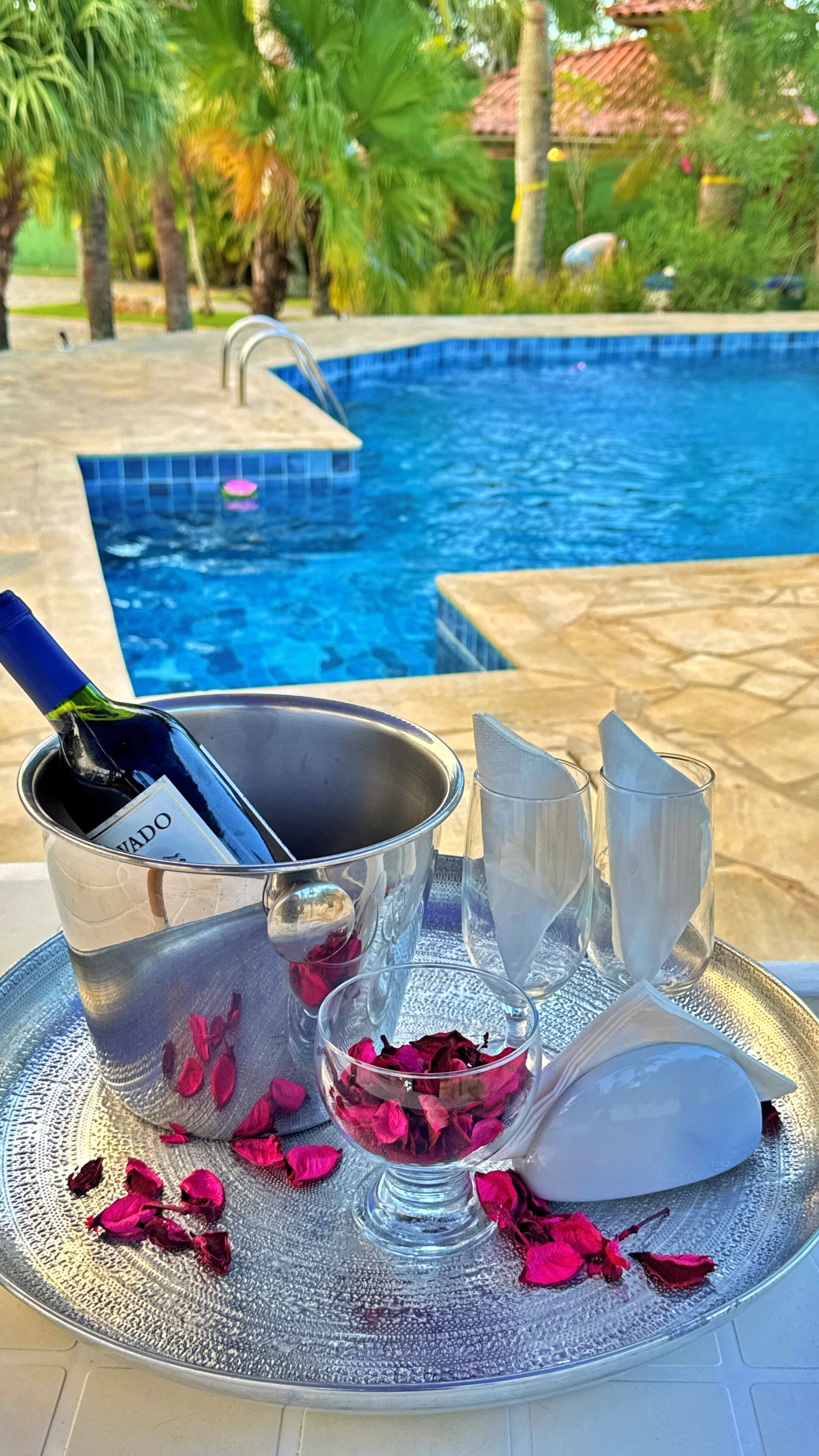 A poolside setting with a metal tray holding a bottle of wine, glasses, and a bowl of rose petals.