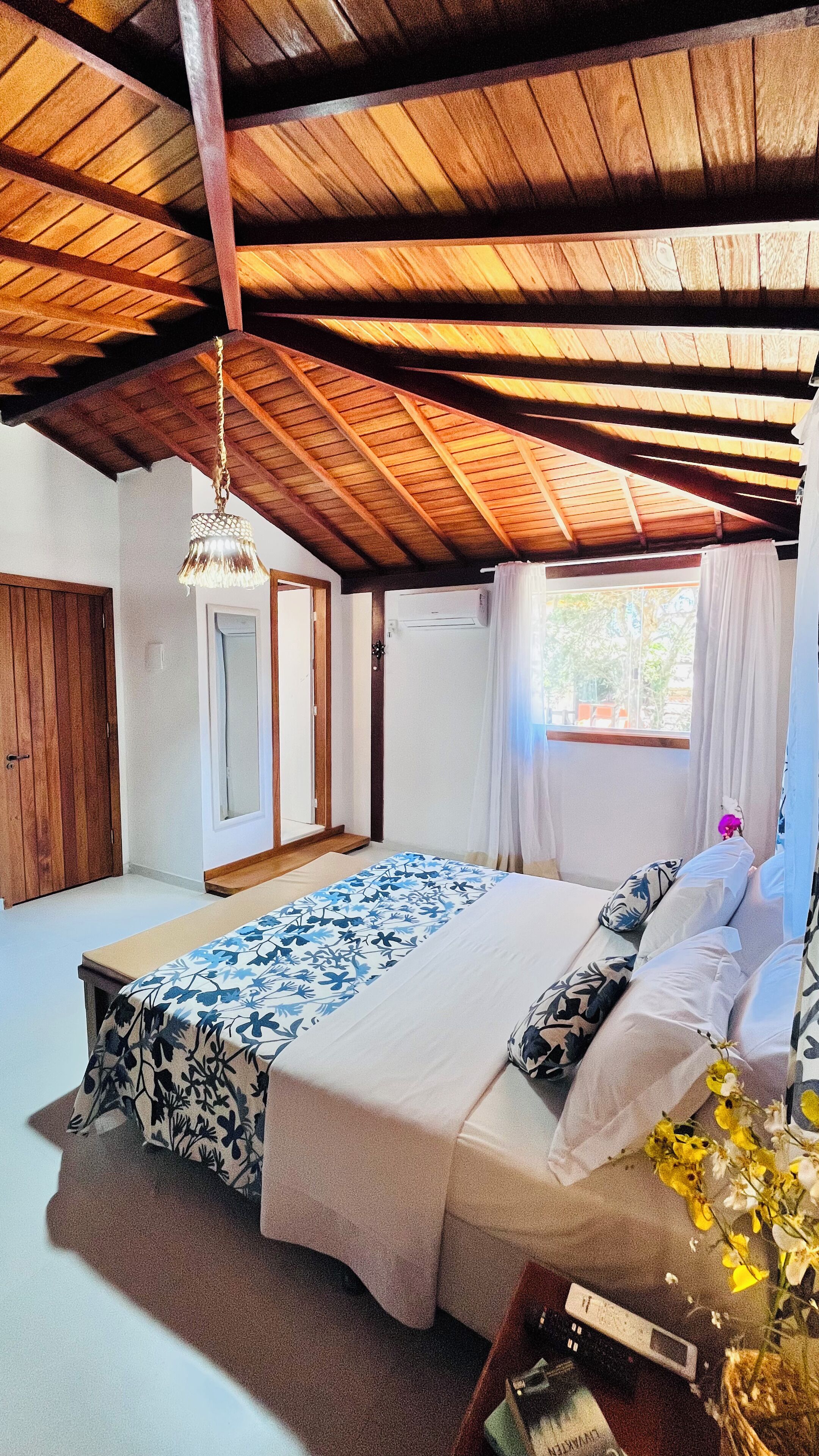 A bedroom with a wooden ceiling, a bed with patterned bedding, a bedside table with a book and a vase of flowers, and a window with curtains.