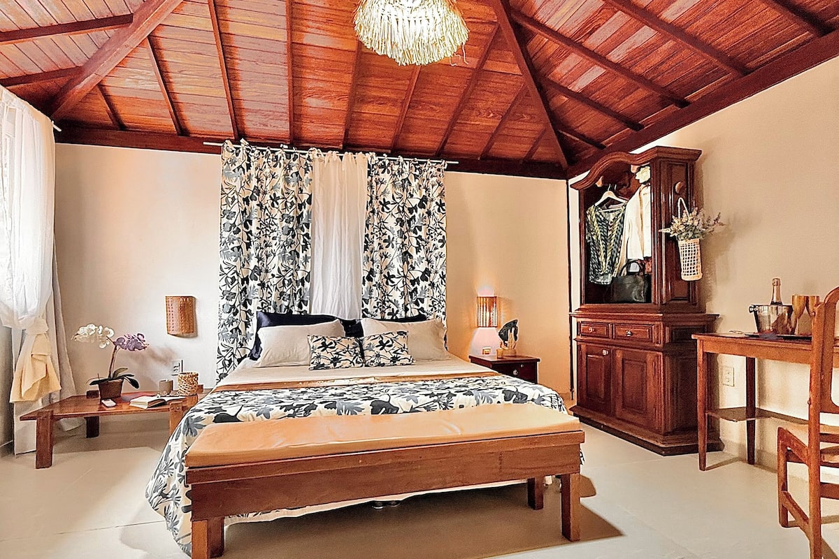 Ein Schlafzimmer mit Holzboden, einem Bett mit Blumenmuster, einer Hängeleuchte, einem Nachttisch, einer Kommode und einem Sessel.