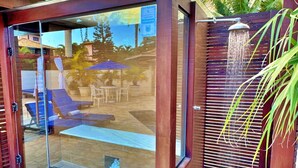 A modern outdoor shower area with a glass enclosure, a wooden deck, and a palm tree. - Villa Baobá (Búzios)
