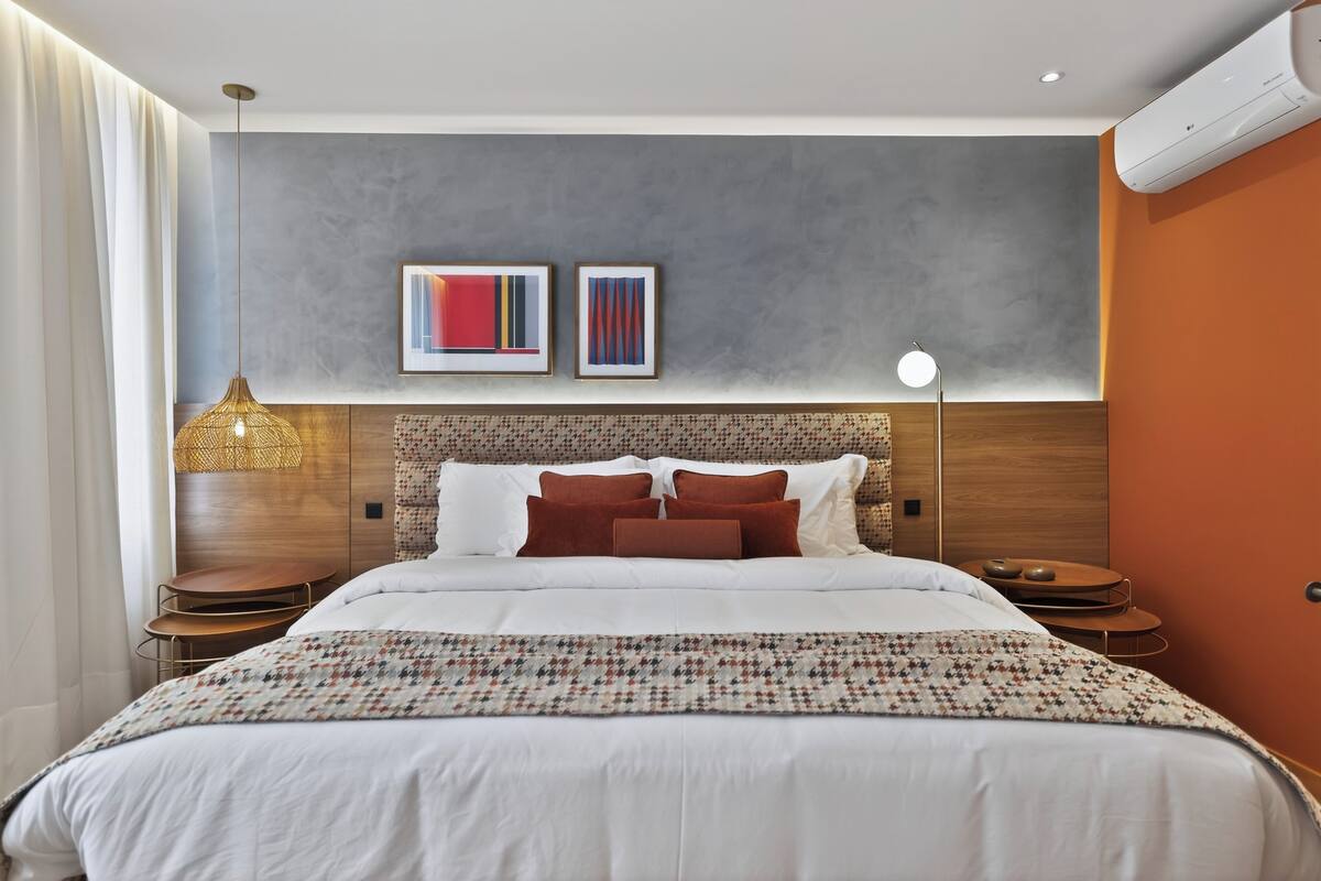 Una habitación de hotel moderna con una cama grande, cabecera con diseño texturizado, dos cuadros en la pared, una lámpara de noche y una mesita con adornos.