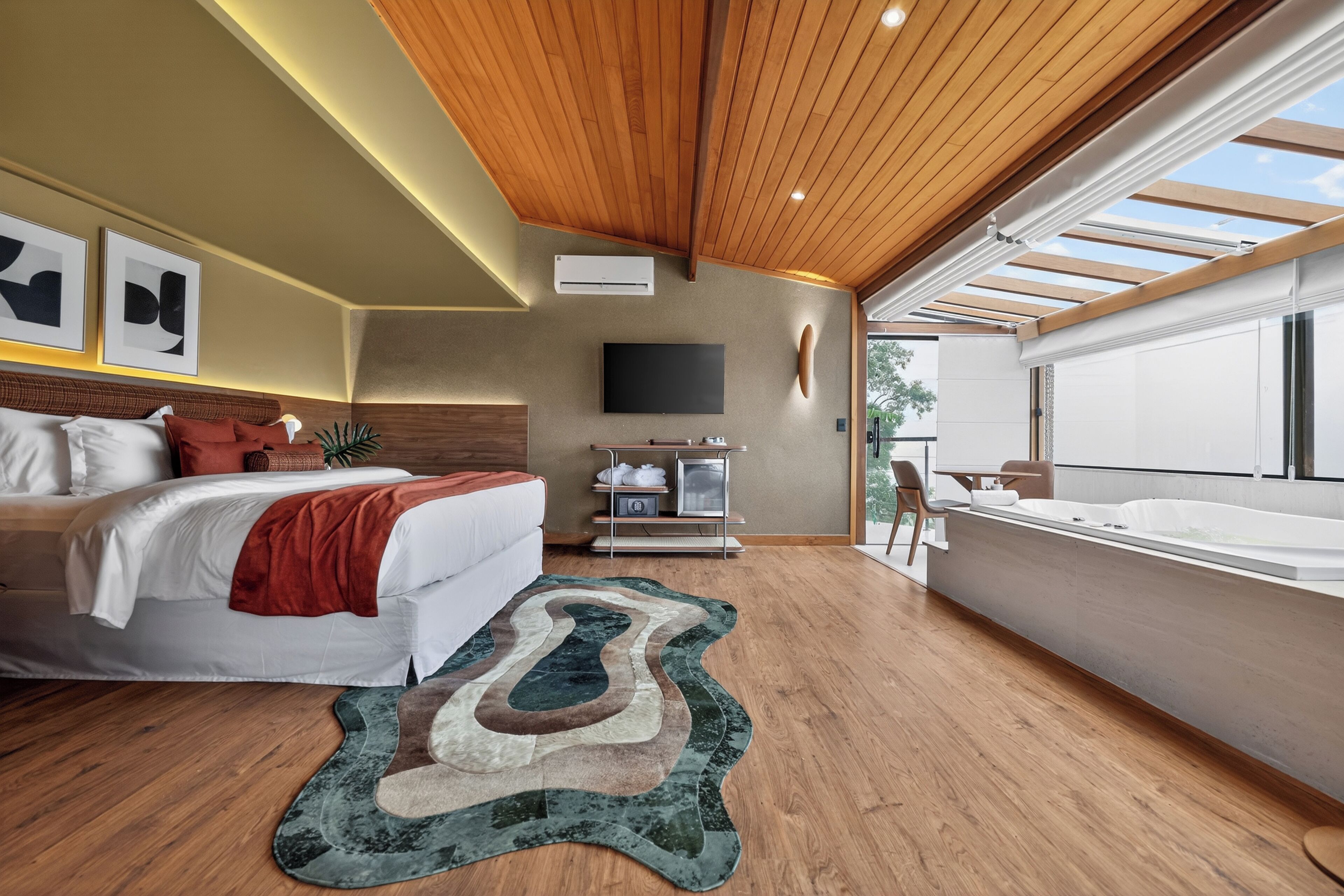 A modern hotel room with a wooden ceiling, a large bed, a flat-screen TV, a bathtub, and a view of the outdoors.