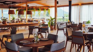 A spacious restaurant with multiple dining tables and chairs, large windows with a view of greenery, and hanging plants. - Siramat Hotel (Petrópolis)