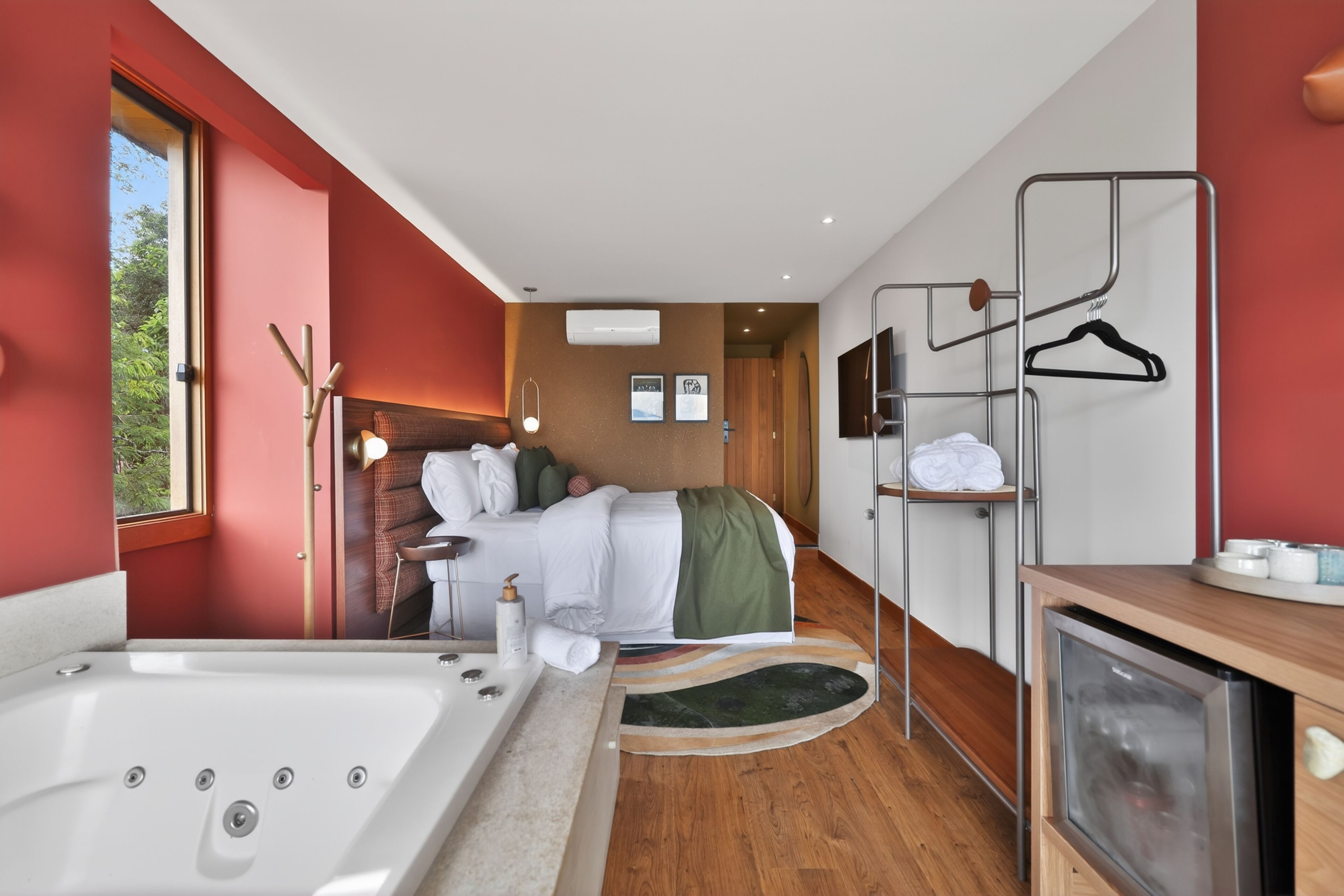 A modern hotel room with a large bathtub, a bed with a green blanket, a wooden floor, and a TV on a wooden cabinet.