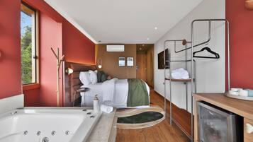 A modern hotel room with a large bathtub, a bed with a green blanket, a wooden floor, and a TV on a wooden cabinet.