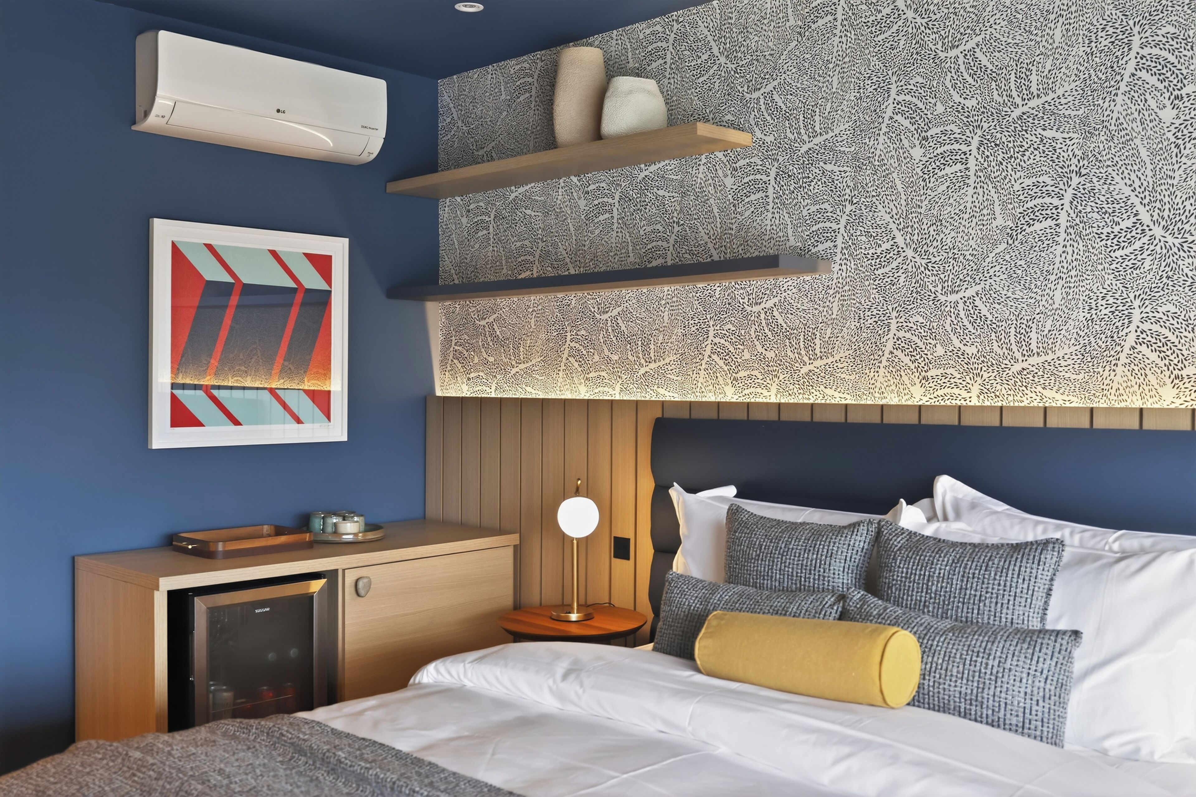 Une chambre d’hôtel moderne avec un mur bleu, un lit avec une literie blanche et grise, une table de chevet en bois, un bureau avec une lampe et un climatiseur fixé au mur.