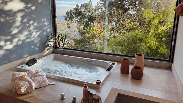 A bathroom with a bathtub, towels, toiletries, and a view of greenery through a window. - Siramat Hotel (Petrópolis)