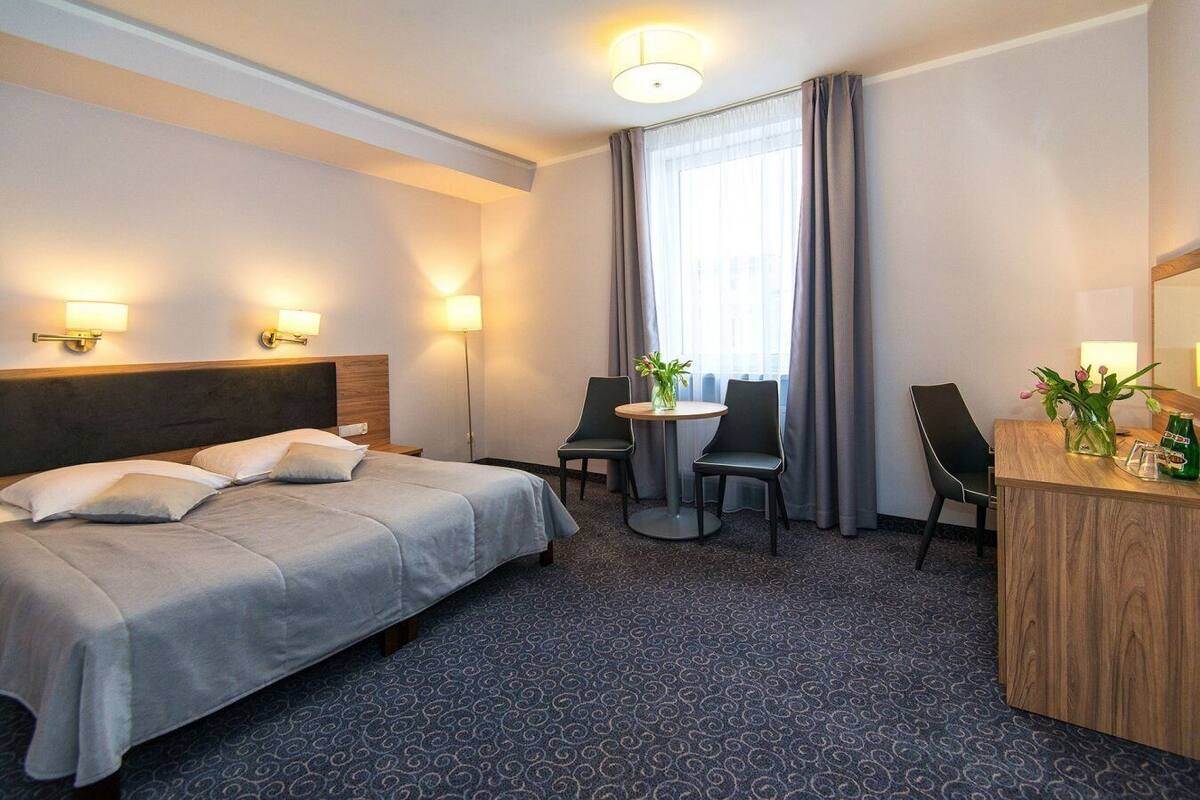 Habitación de hotel con cama, dos sillas, una mesita, escritorio y ventana con cortinas.