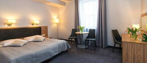 A hotel room with a bed, two chairs, a small table, a desk, and a window with curtains.