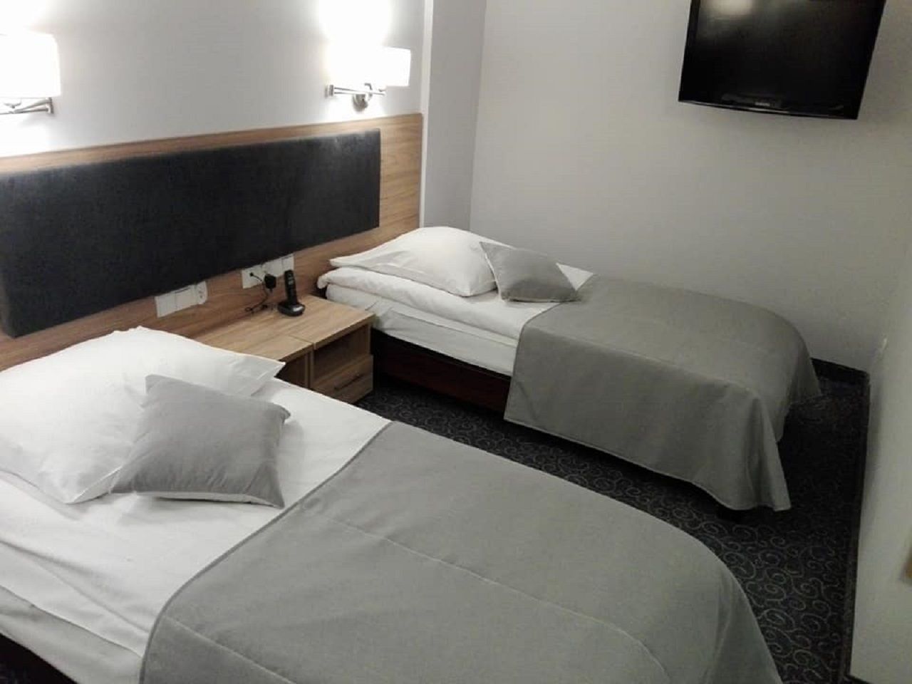 Habitación de hotel con dos camas individuales, un cabecero de madera, una mesita de noche, un televisor y una alfombra estampada.