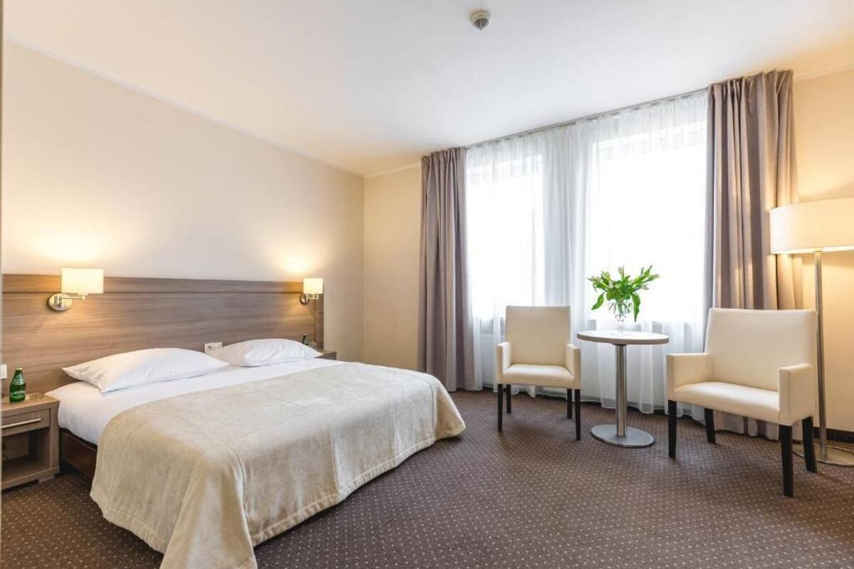 Habitación de hotel con una cama grande, dos mesitas de noche, una mesa redonda con dos sillas, una planta en maceta y una ventana con cortinas.