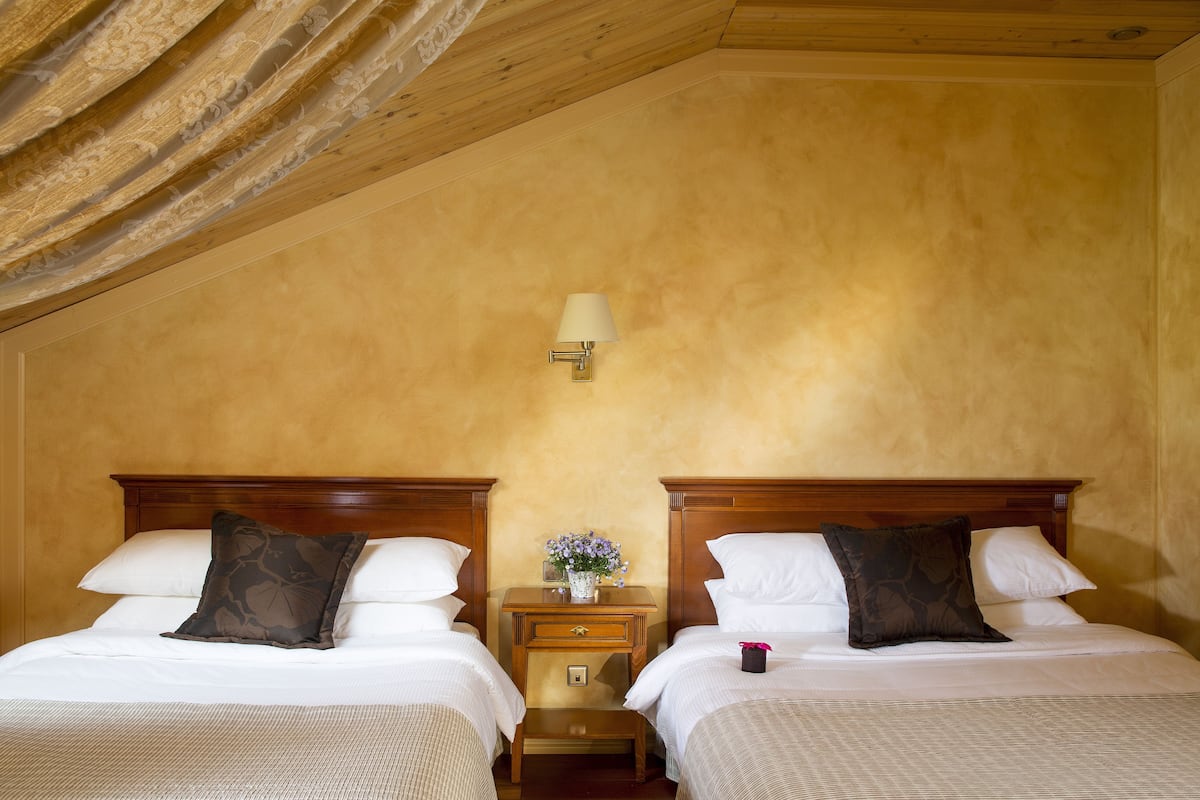 A room with two beds, wooden headboards, a nightstand with a lamp, and a vase with flowers.