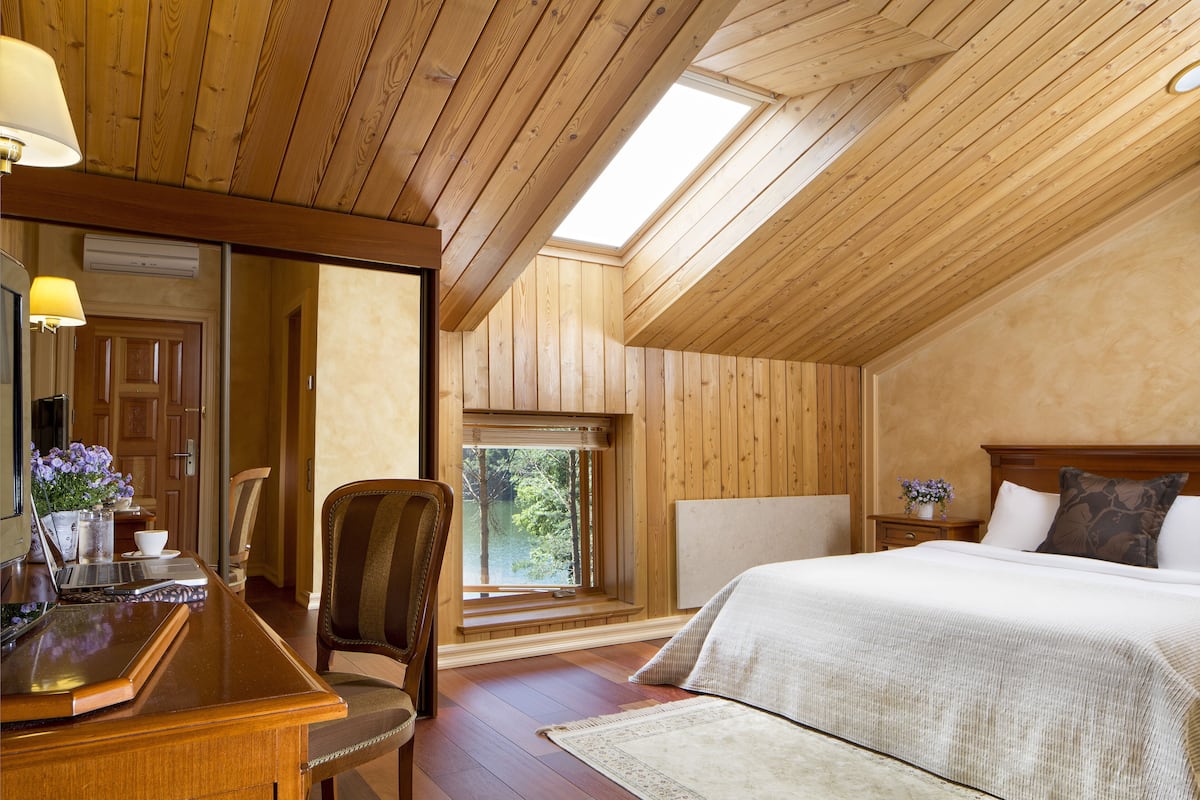 A bedroom with a wooden ceiling, a bed, a desk with a laptop, and a window with a view of trees.