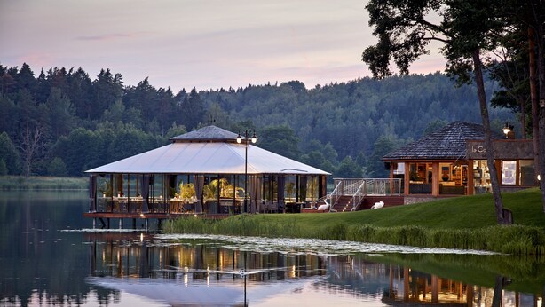 Ein Seehaus mit einem wintergartenartigen Essbereich, einem separaten Gästehaus und einem Spiegelbild im Wasser.