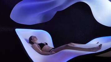 A woman lying on a curved, illuminated lounge chair in a dark room with a starry background.