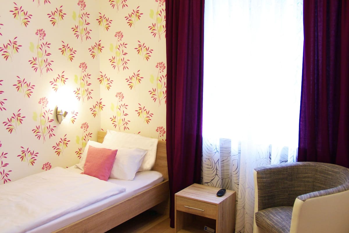 Un dormitorio con cama, mesita de noche, silla y ventana con cortinas.