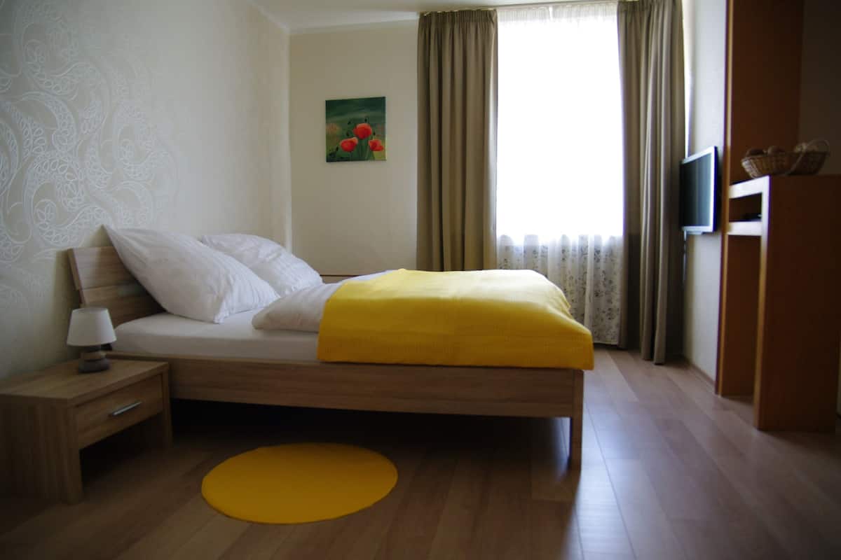 Un dormitorio con cama, una mesita de noche, una alfombra amarilla, un cuadro en la pared y un televisor en la pared.