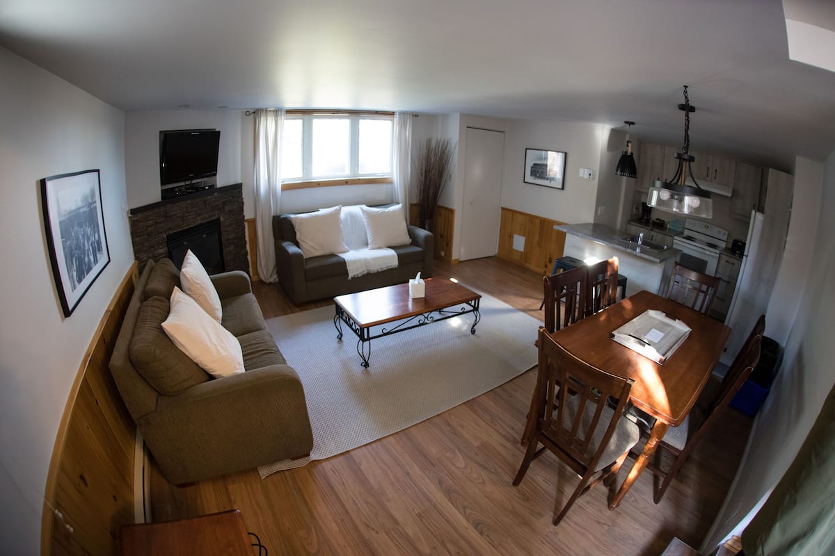 Ein Wohnzimmer mit einer Couch, einem Couchtisch und einem Fernseher. Es gibt einen Kamin, einen Essbereich mit Stühlen und eine Küche mit Schränken und Geräten.