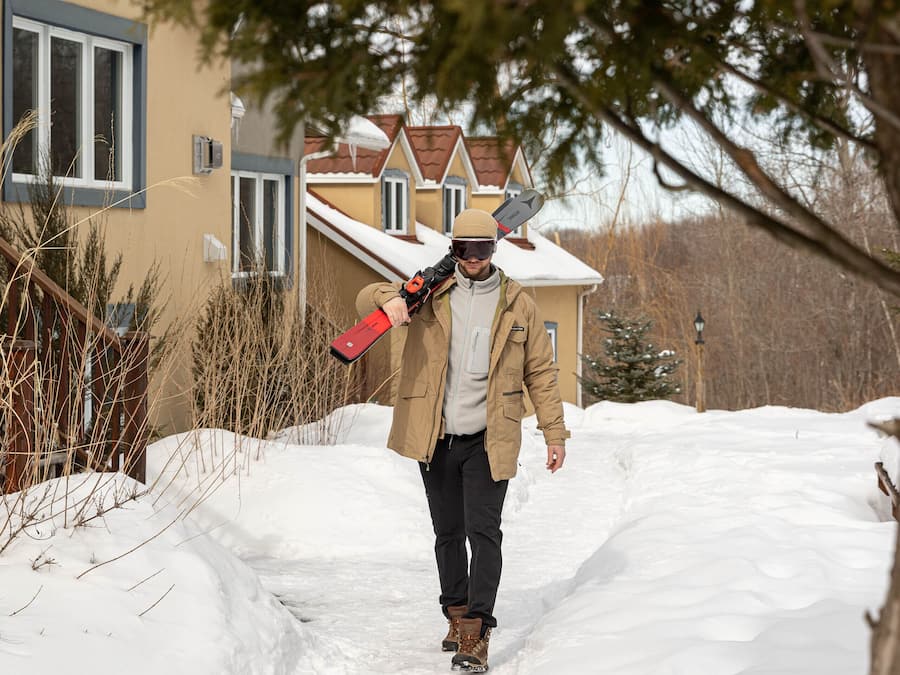 Una persona caminando por un sendero cubierto de nieve hacia una casa con techo nevado y una ventana de marco blanco.
