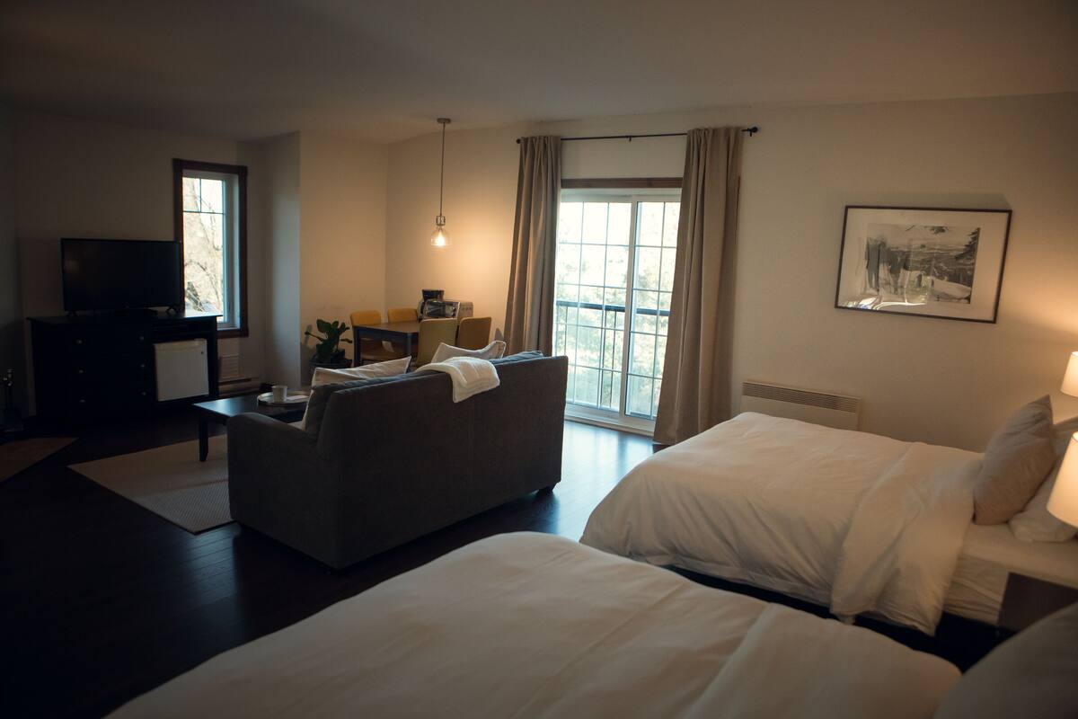 Ein Hotelzimmer mit einer Sitzecke, zwei Betten, einem Fernseher, einem Schreibtisch und einem Fenster mit Vorhängen.