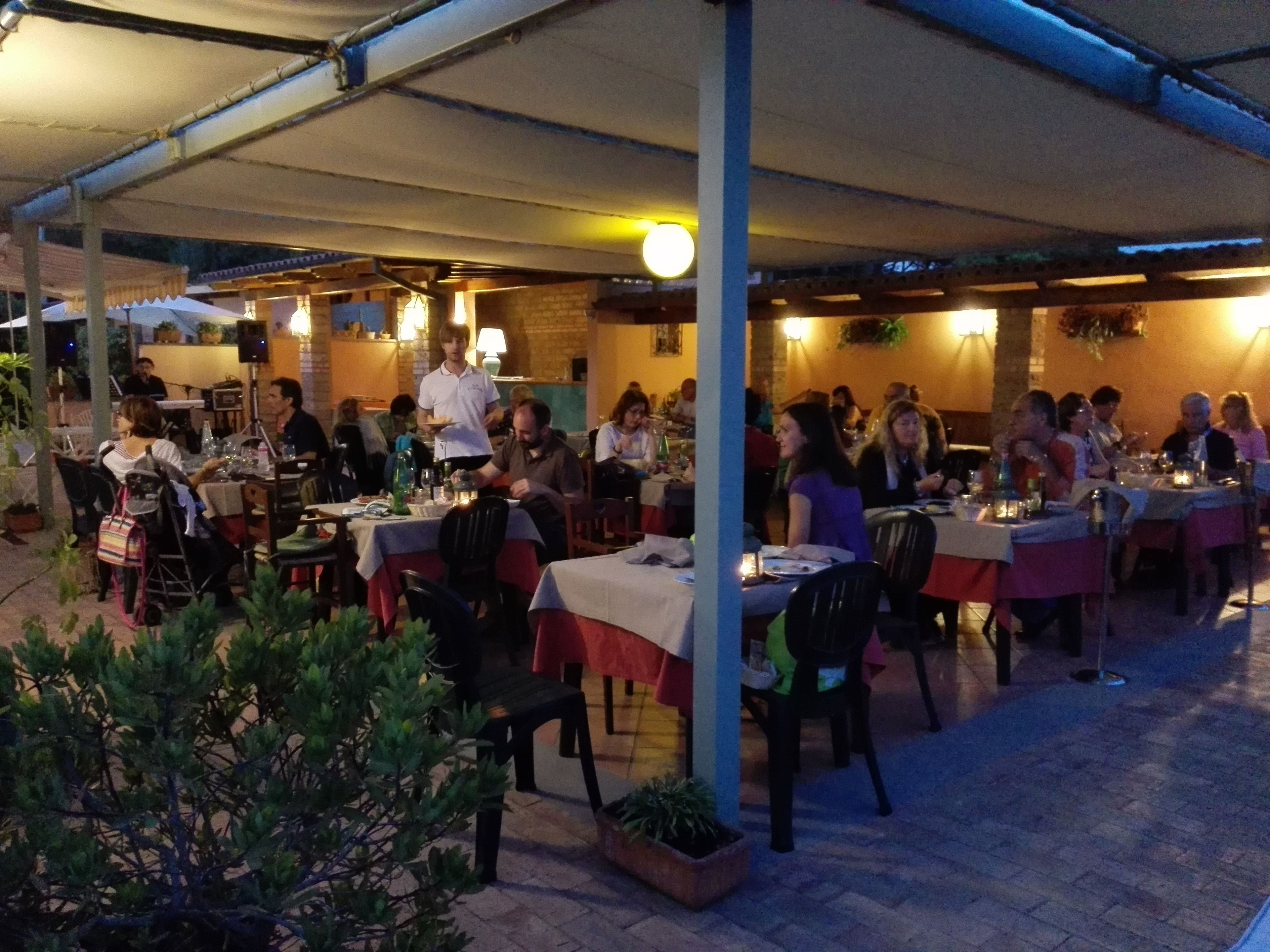 An outdoor dining area with tables and chairs, covered by a roof, and surrounded by potted plants.