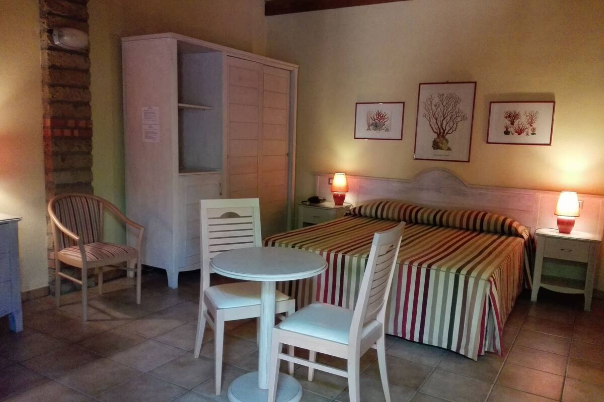 Una camera d'albergo con un letto, due poltrone, un tavolino, un armadio e due lampade ai comodini.