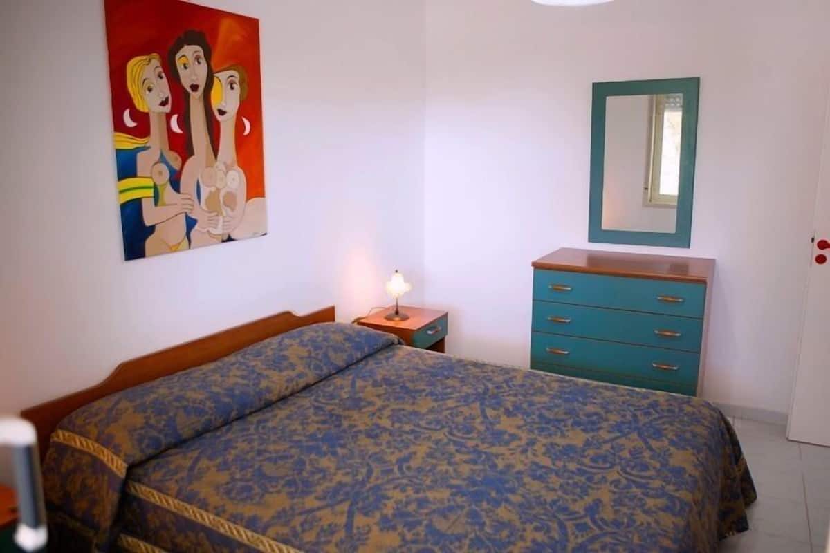 A bedroom with a bed, a dresser, a nightstand, a painting, and a mirror.