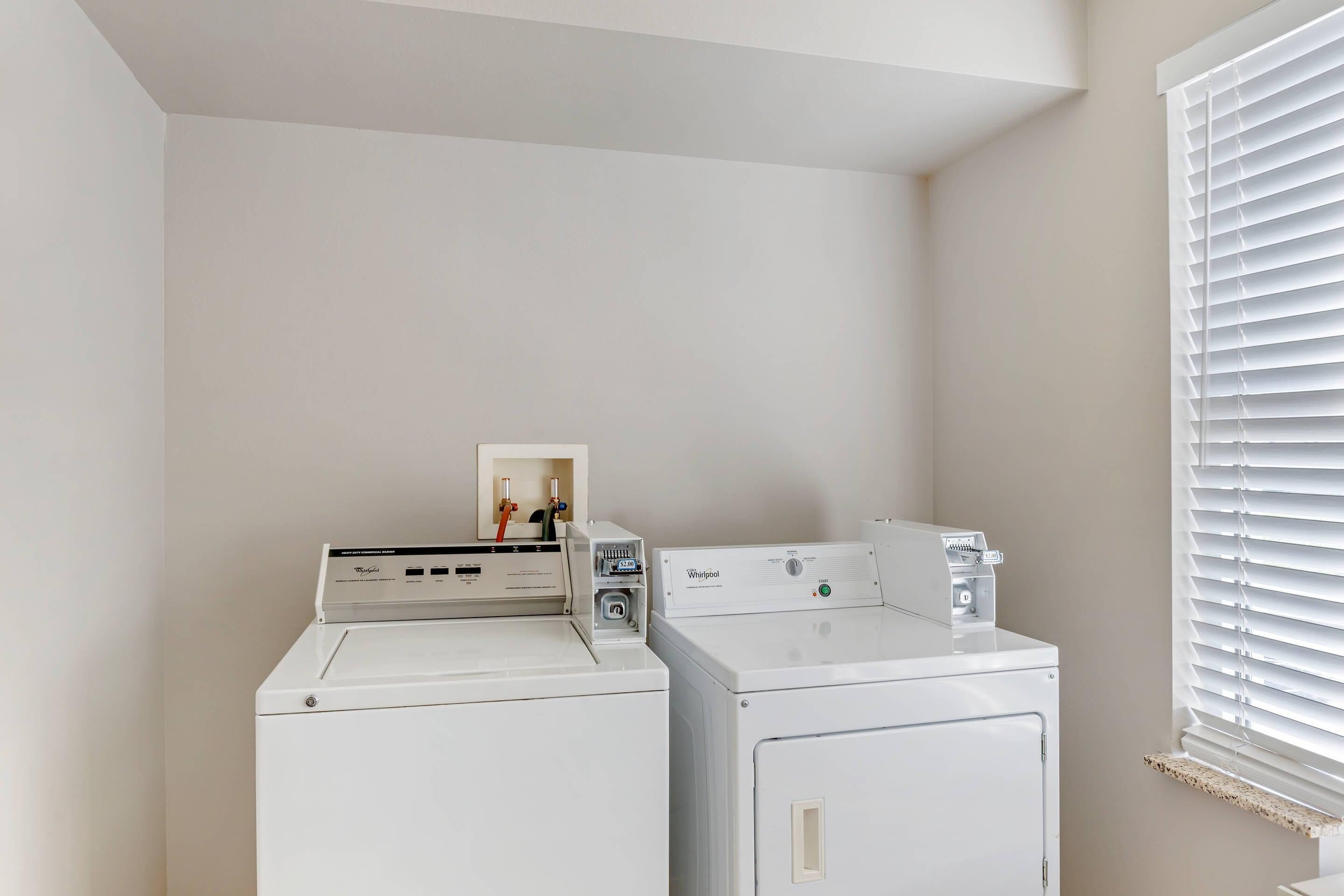 Uma lavanderia com duas máquinas de lavar e secar Whirlpool brancas, uma prateleira pequena com um porta-retrato, e uma janela com persianas.
