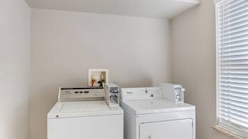 Uma lavanderia com duas máquinas de lavar e secar Whirlpool brancas, uma prateleira pequena com um porta-retrato, e uma janela com persianas.